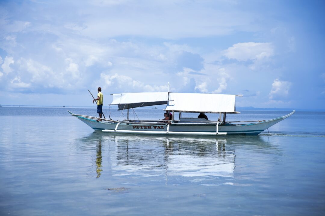 A small white boat with a man stood at the front propelling the boat with an oar. There are passengers sat in the boat underneath a roof.