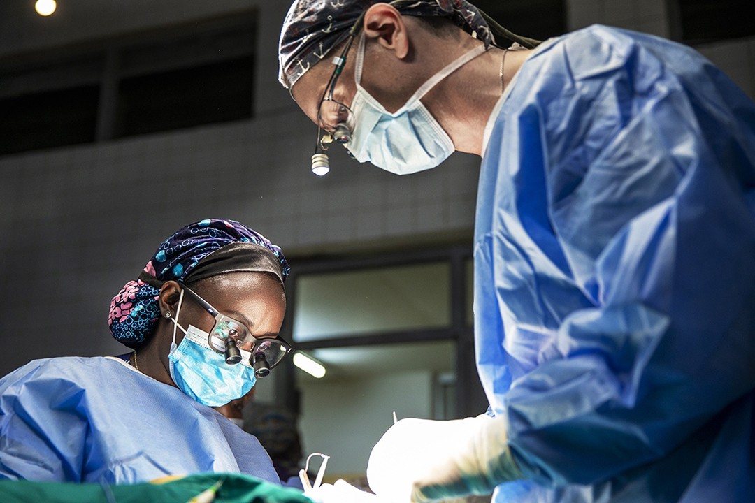 Surgeon wearing gown and face mask operates on an unseen person. Photo by Jorgen Hildebrandt.