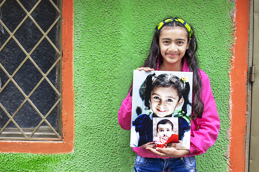 Teenage girl stands in front of a bright green house holding images of herself as a younger child and as a baby.
