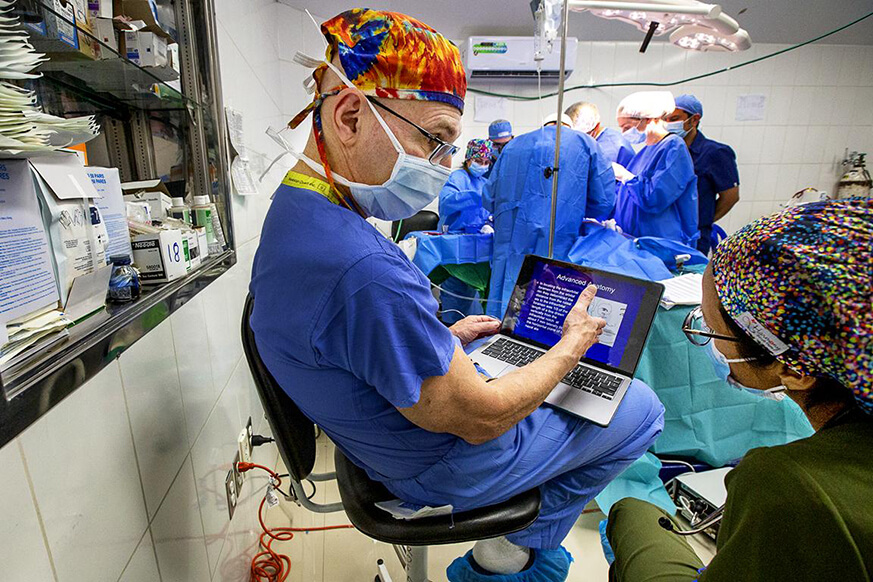 Anesthesiologist is sat on a chair with a laptop on his knee, talking to his mentee. In the background, surgery is taking place.
