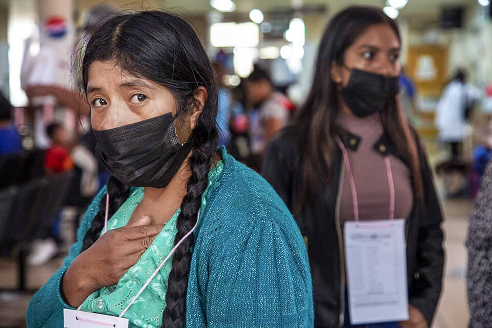 Patient Paulina, wearing a black face mask and green top and cardigan, stands in a waiting area, placing her hand on her heart.