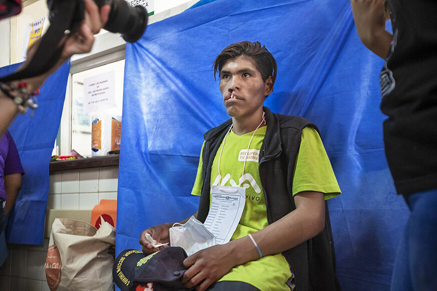 Teofilo, wearing a bright green tshirt and a black sleeveless jacket, and holding a black baseball cap in his hands, sits down on a chair.