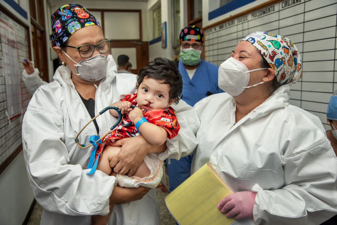 10-month-old Juan Elias, wearing a red patterned gown, is carried to the operating room by Drs. Carla Garcia and Silvia Ramos, both wearing white coats, face masks and patterned surgical caps. 