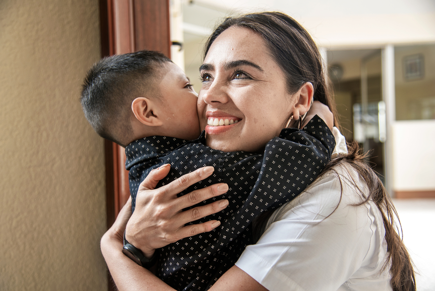 Bea Vidal, with long brown hear and wearing a white shirt, embraces a young male patient in the care centre. He has short brown hair and is wearing a black shirt with a small white flower pattern.