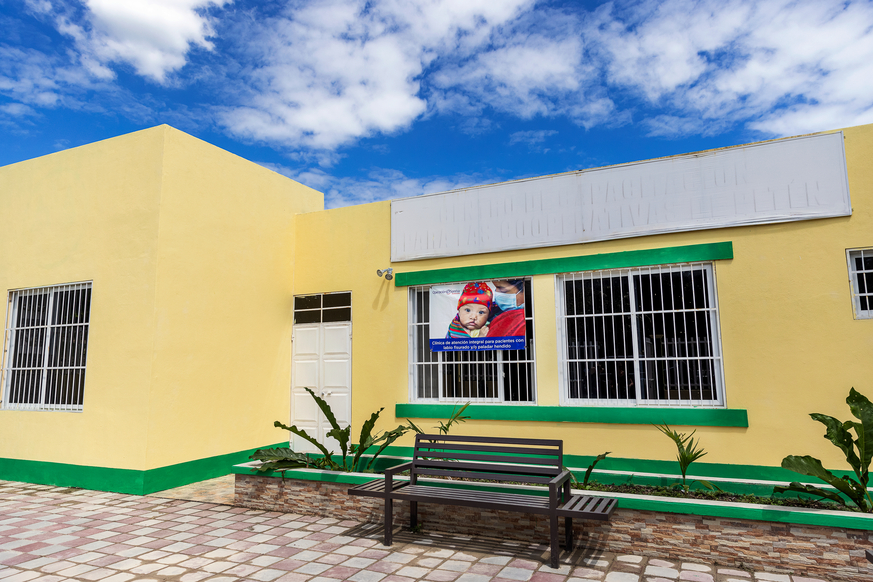 The Petén center is a single storey building. It is painted yellow, with window sills and the bottom of the building painted green. There are plants in front of it, with a brick path leading to a white door. 