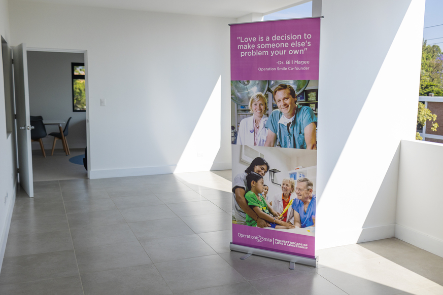 An empty room in the comprehensive care centre. It has white walls and a beige tiled floor. There is a banner in the middle of the room with pictures and a quote: "Love is a decision to make someone else's problem your own", Dr Bill Magee, Operation Smile Co-founder 