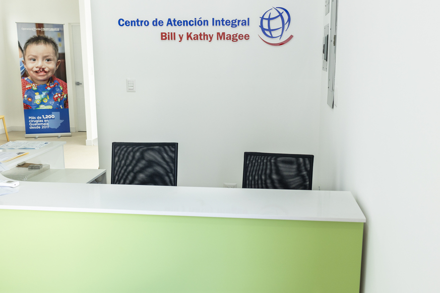 A green reception desk, with two empty chairs sat behind. On the wall is a sign that says 'Centro de Atencion Integral Bill y Kathy Magee' (Bill and Kathy Magee Comprehensive Care Centre)