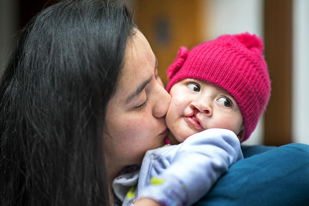 6 month old Luciana before her cleft surgery. She is wearing a pink knitted hat and a lilac cardigan, and her mum kisses her on the cheek.
