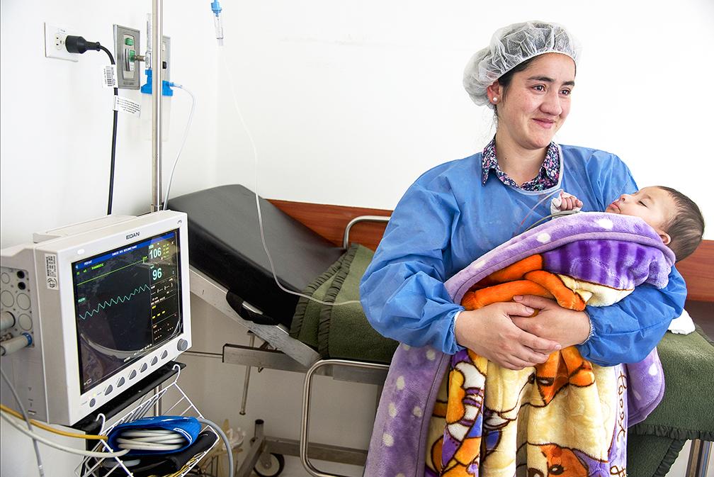 Luciana is held by her mother after her cleft surgery. Her mother is wearing surgical hat and gown, and Luciana is asleep, wrapped in a blanket.