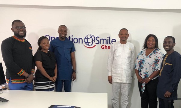 Six people from Operation Smile and the Ghana Ministry of Health stand in front of a white wall with the Operation Smile Ghana logo.