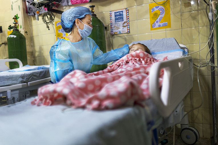 Ana strokes her daughter Zoe's head, who is lying on a hospital bed under a pink spotted blanket.