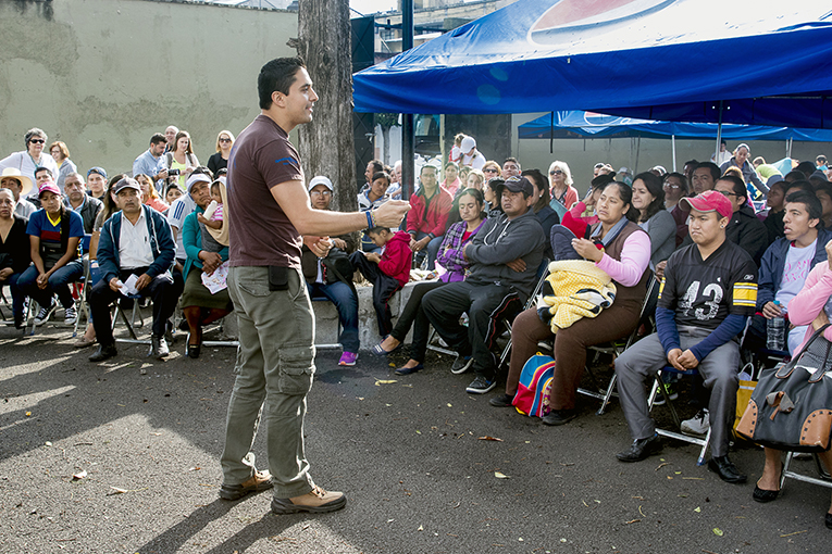 A man stands and speaks in front of a seated outdoor audience, who are attentively listening. The crowd includes people of various ages, some holding notebooks and blankets. A blue canopy provides shade for some audience members. Trees and buildings are in the background.