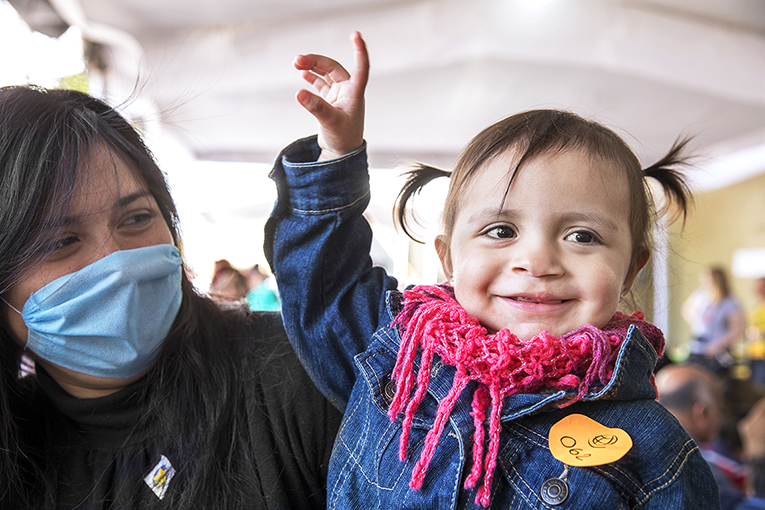 A young child with pigtails and a pink scarf smiles and raises a hand, while being held by a woman with long dark hair, wearing a blue face mask. The child wears a denim jacket with a yellow sticker. The scene appears to be in a busy indoor setting.
