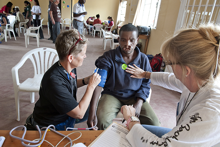 Two medical professionals examine a seated patient’s blood pressure in a clinic. The patient has a cleft lip. Other medical personnel and patients are visible in the background. White plastic chairs and a spacious room provide a setting for the scene.