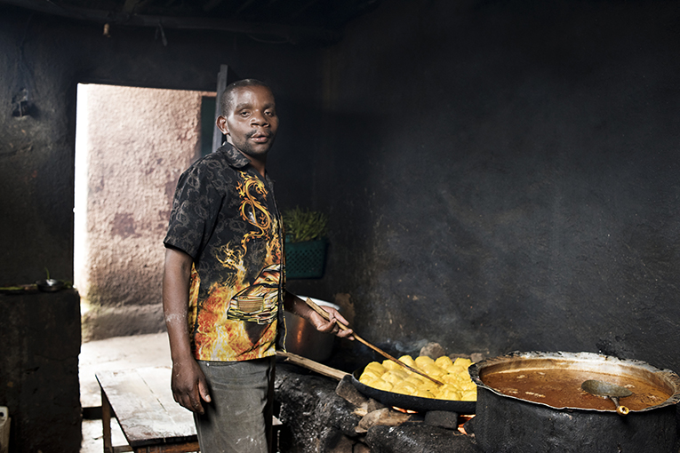 Enok stands in a dimly lit kitchen in front of a large pan filled with food, turning it with a utensil. He wears a patterned short-sleeve shirt and looks at the camera. An open doorway behind him reveals some natural light entering the room.