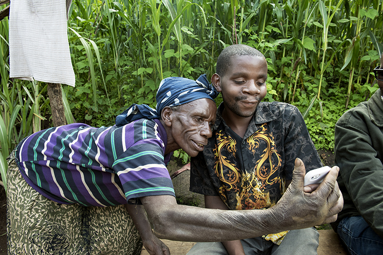 An elderly woman wearing a striped shirt and patterned headscarf leans close to a young man holding a smartphone. They both smile as they look at the phone screen, likely taking a selfie. They are outdoors with lush green vegetation in the background.