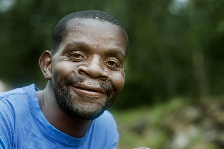 A man with a short beard, wearing a blue shirt, smiles warmly at the camera. He appears to be outdoors with blurred greenery in the background.