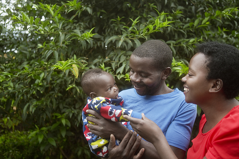 A smiling man in a blue shirt holds a baby dressed in a colorful outfit, while a smiling woman in a red shirt touches the baby's hand. They are standing outdoors, with lush green foliage in the background.
