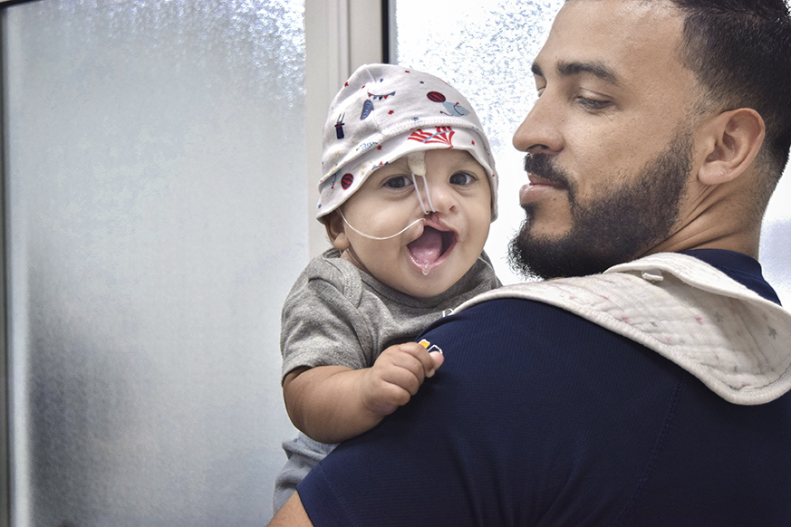 A man holds a smiling baby wearing a patterned hat and a nasal feeding tube. They stand near a window with frosted glass. The man looks at the baby with affection.