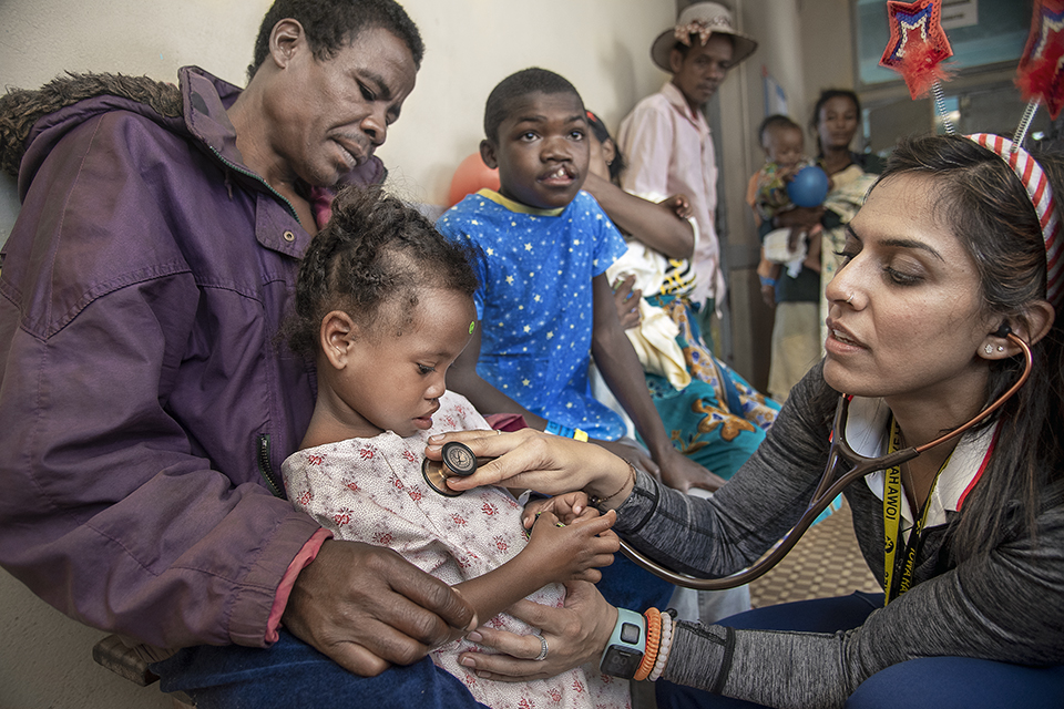 A healthcare worker uses a stethoscope to examine a young child sitting on an adult's lap in a clinic. Other children and adults wait in the background, some in colorful clothing. The setting appears to be a busy healthcare facility.