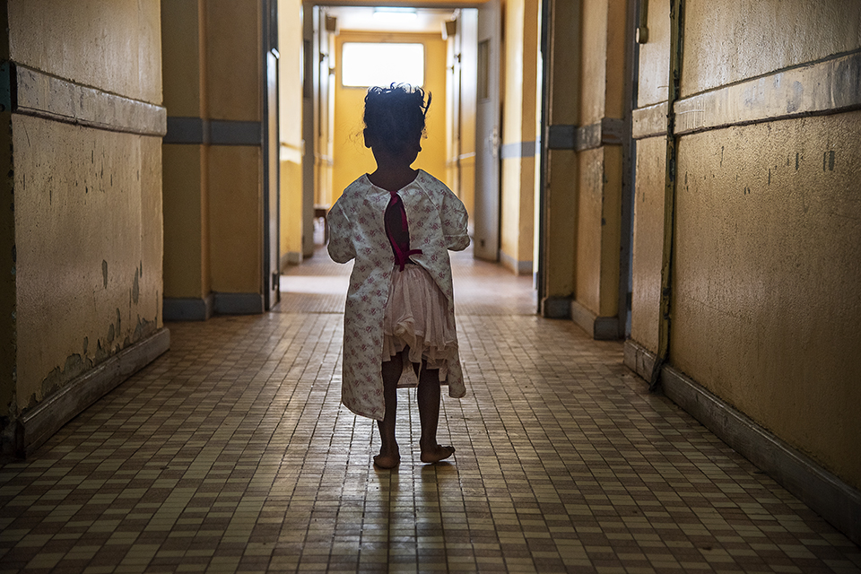 A child in a hospital gown stands alone in a dimly lit corridor of an old building. The hallway has yellow walls and a patterned tile floor, adding to the somber atmosphere. Light shines through an open doorway at the end of the hall.