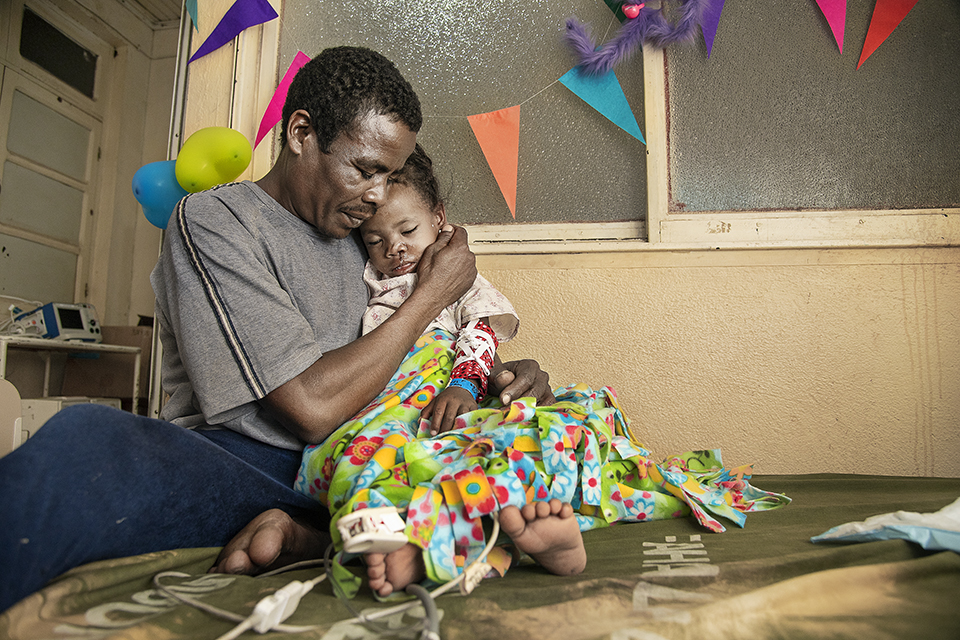 A man lovingly holds a child wrapped in a colorful blanket, sitting on a bed. The room is decorated with colorful triangular flags. Medical equipment is visible in the background, suggesting a hospital setting. The child's eyes are closed as they rest.