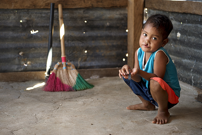 A young child in a blue tank top and shorts crouches on a concrete floor in a rustic room. Behind, there are colorful brooms leaning against a corrugated metal wall. The child looks at the camera with a curious expression.