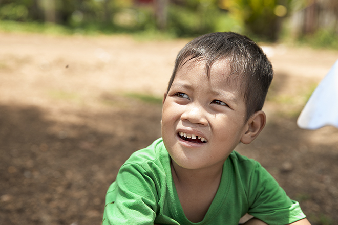 A young boy with short hair and wearing a green shirt looks off to the side with an expression of curiosity or confusion. The background is slightly blurred, showing a sunny, outdoor scene with brown soil and green foliage.