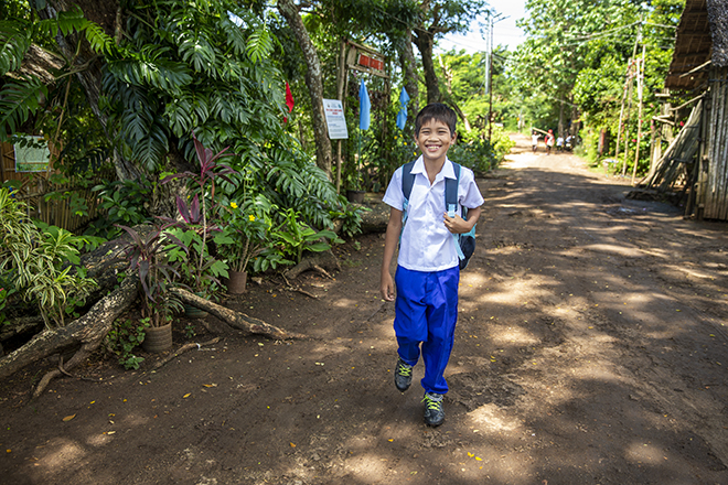 A young boy in a white shirt and blue pants walks along a dirt road, smiling and carrying a backpack. He is surrounded by green trees and vegetation. The sunny setting suggests a rural area.