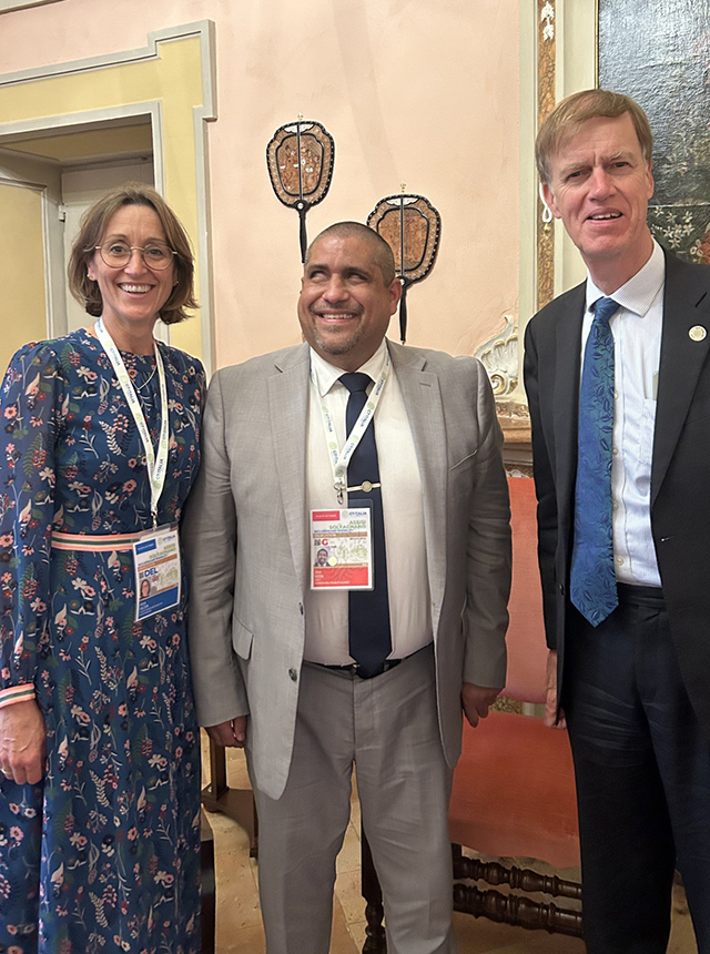 Three individuals smile for a photo indoors. The woman on the left wears a patterned dress, the man in the center is in a light suit, and the man on the right wears a dark suit with a blue tie. They stand in a room with ornate decor.