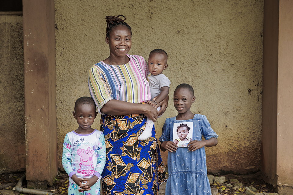 A woman smiles while holding a baby in the Philippines. Two children stand beside her; one holds a photo of a child with cleft lip. The group is against a textured wall. The woman is wearing a colorful striped top and patterned skirt, and the children are in casual attire.