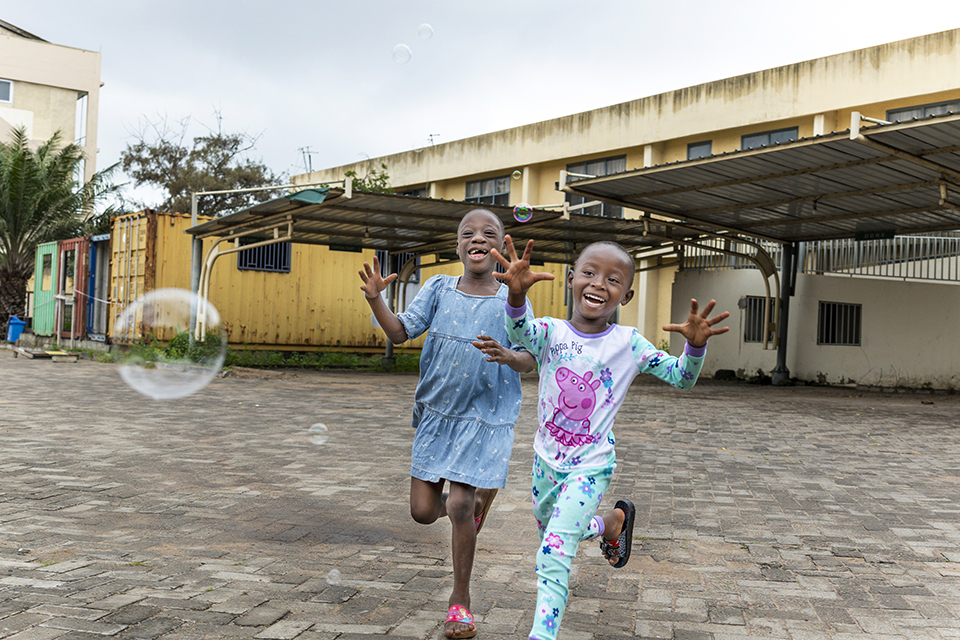 Two children joyfully run on a paved surface in the Philippines, reaching for soap bubbles. The first child wears a light blue dress, and the second dons a white outfit with colorful designs. A covered parking area and buildings are visible in the background.