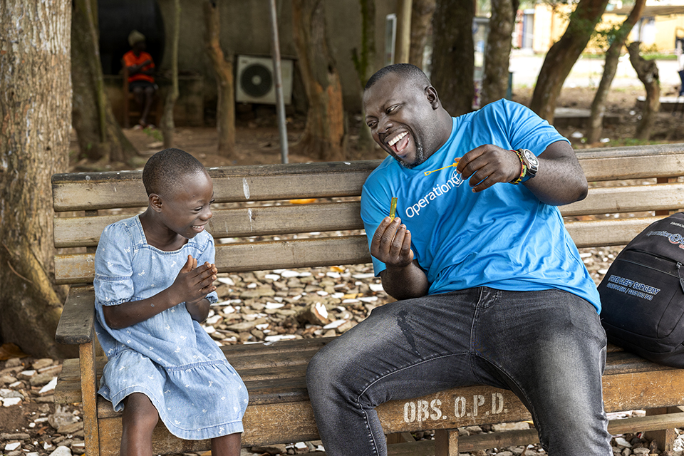 A man and a young girl share a joyful moment on a wooden bench in Ghana. The man, wearing a blue T-shirt, holds a yellow candy while the girl, dressed in a light blue dress, laughs heartily. Trees and a blurred background add to their blissful scene.