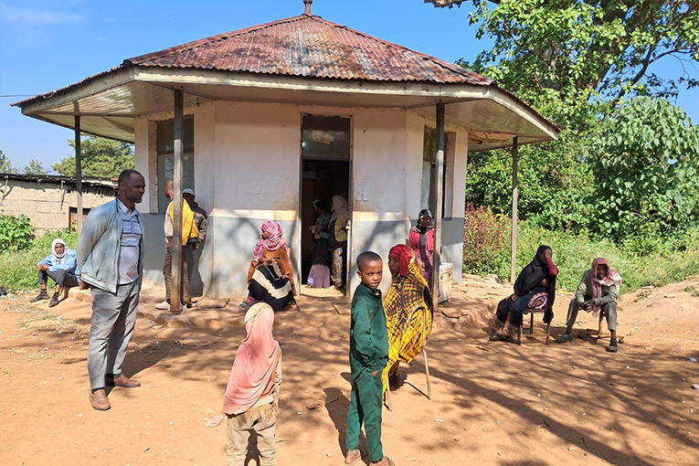 A group of people, including children, gather outside a small, round building with a rusty metal roof. Some are sitting while others are standing on a dirt path, with trees and blue sky in the background.