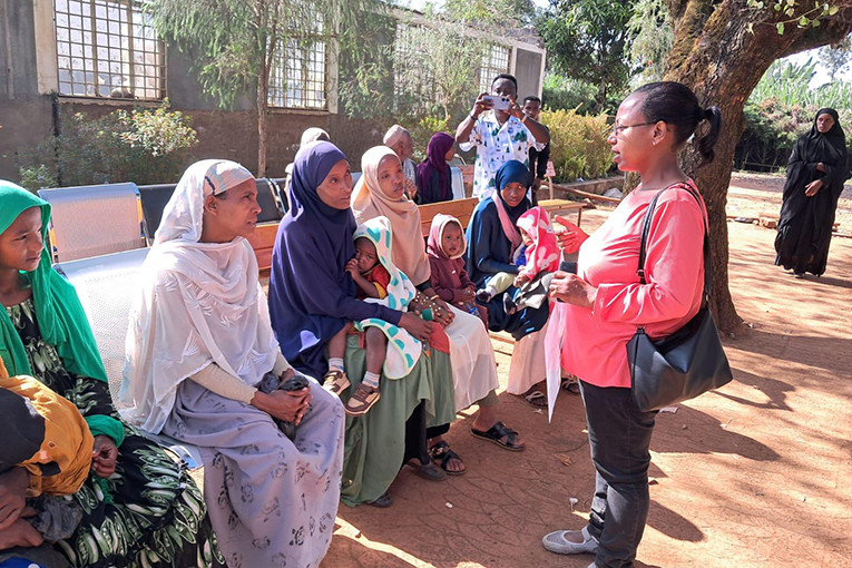 A woman in a red shirt talks to a group of seated women and children outdoors. The women are wearing colorful headscarves. Trees and a building are in the background. Some women hold infants, and the setting appears social and relaxed.