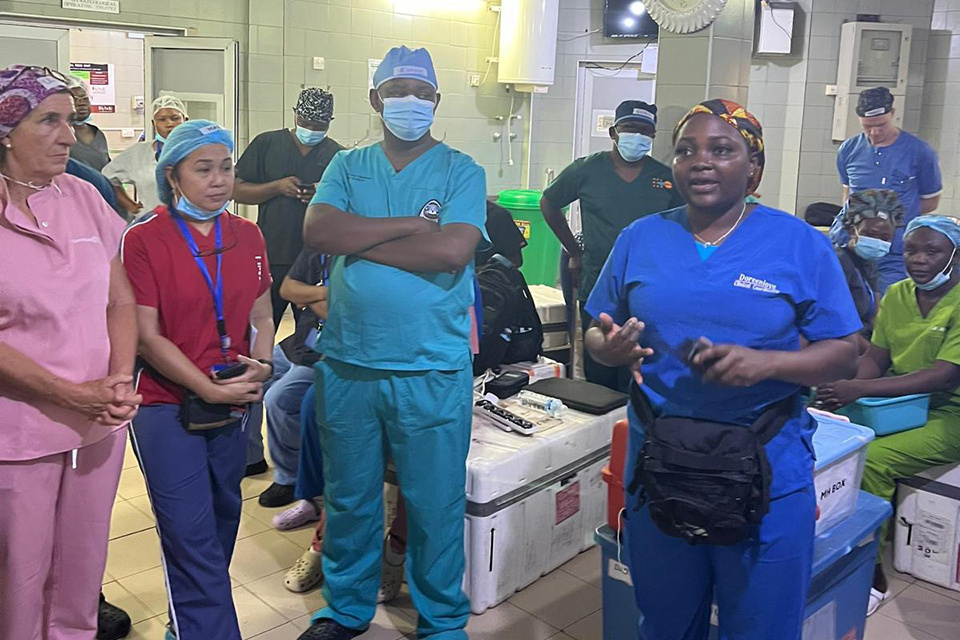 A group of medical professionals in scrubs and masks stand and sit together in a hospital room, listening attentively as one woman in blue scrubs speaks to them. Medical equipment and supplies are visible in the background.