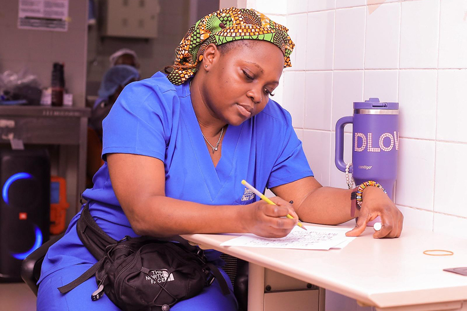 A woman in blue scrubs and a patterned headscarf sits at a table writing on a piece of paper. A large water bottle and a black waist bag are on the table beside her. She appears focused on her work.