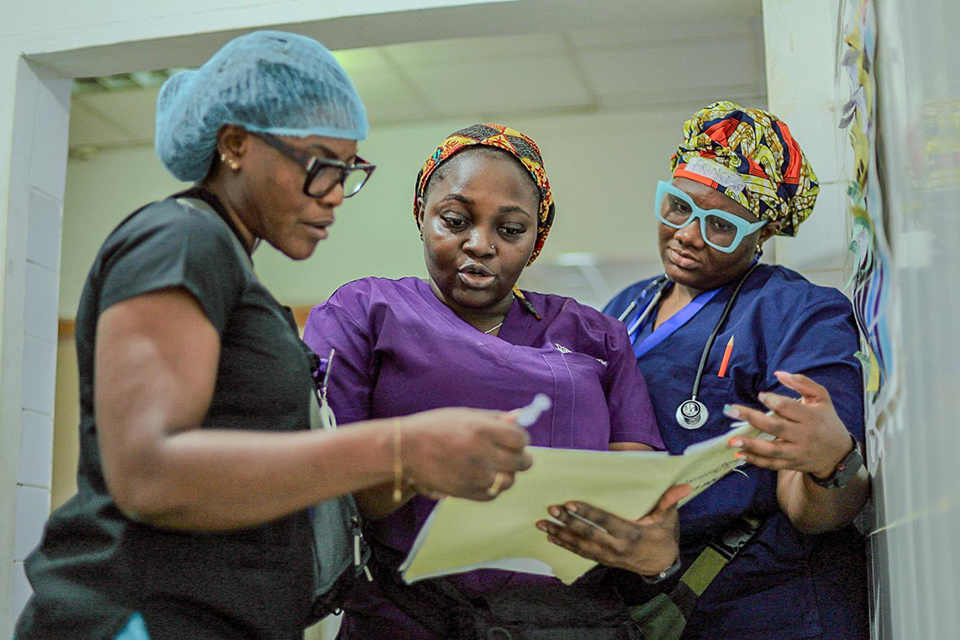 Three healthcare professionals in colorful scrubs and head coverings stand together, reviewing documents or medical charts in a well-lit hospital hallway.