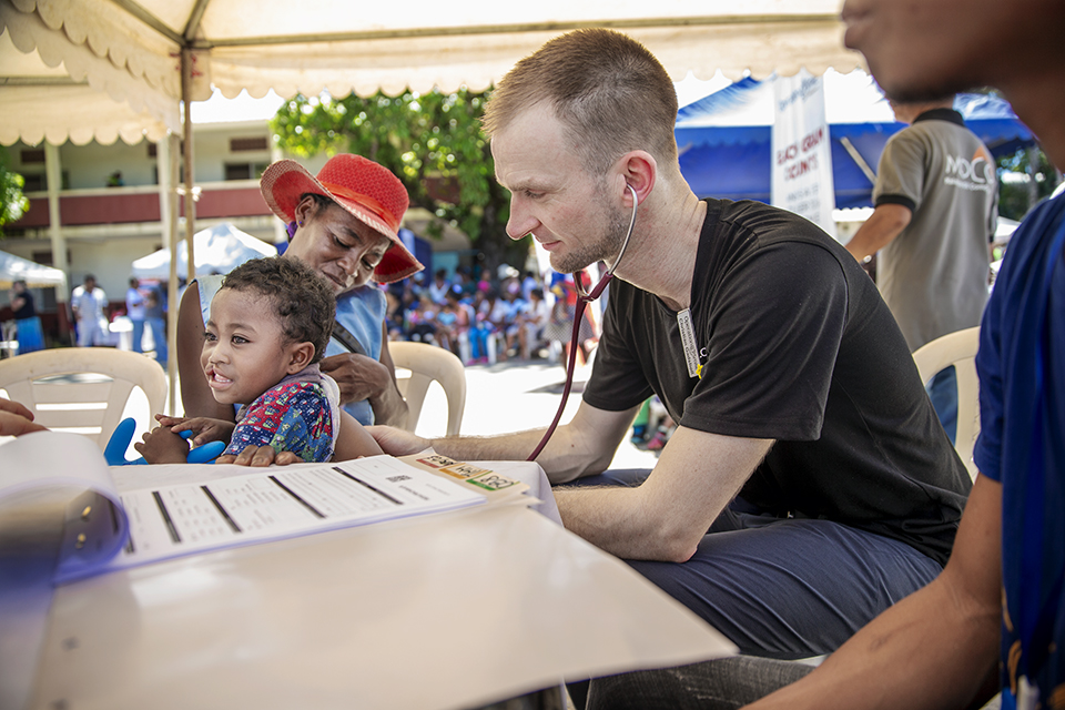A healthcare worker uses a stethoscope to check a young child’s heartbeat at an outdoor medical clinic, while the child sits on an adult’s lap and smiles. Other people and tents are visible in the background.