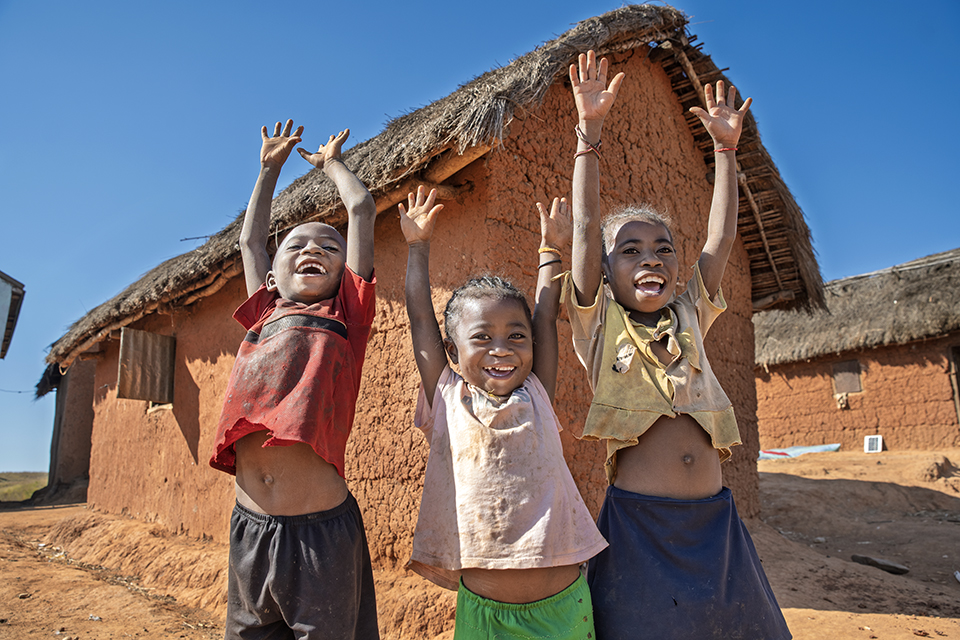 Three smiling children with raised arms stand in front of traditional thatched-roof mud houses under a clear blue sky. They appear joyful and playful, with their shirts lifted, revealing their midriffs.