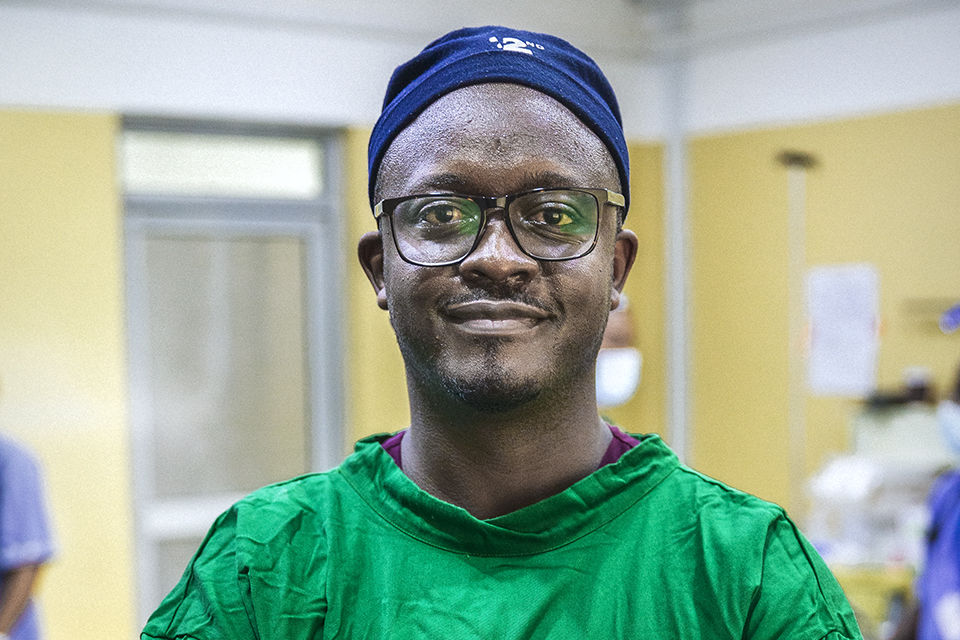 A man wearing glasses, a blue surgical cap, and a green surgical gown stands in a hospital room, looking at the camera with a slight smile. The background is blurred, showing medical equipment and another person.