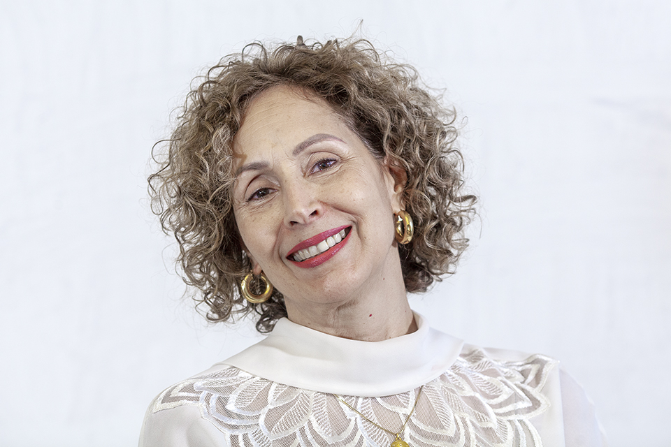 Woman with curly hair smiling, wearing a white lace top and gold hoop earrings, against a light background.