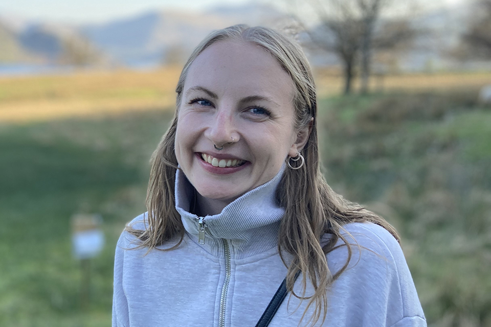 A young woman with light brown hair and hoop earrings smiles outdoors, wearing a light gray zip-up sweater. The background features a grassy field, trees, and blurred mountains.