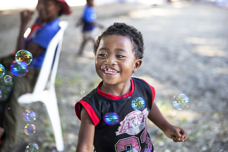A young child smiles joyfully while playing with bubbles outdoors. The child wears a black and red shirt and is surrounded by floating bubbles, with blurred people and a white chair in the background.