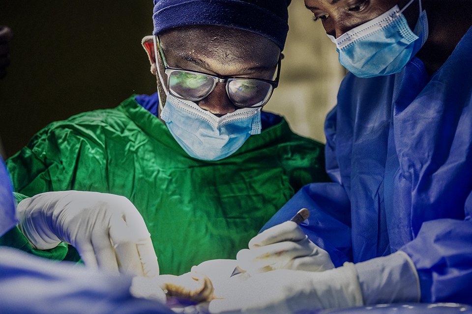 Two surgeons wearing masks, glasses, and surgical gowns carefully perform an operation under bright light, focused on the procedure with gloved hands working together.