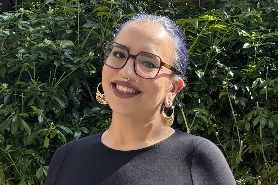 A person with glasses, purple hair, and large hoop earrings smiles outdoors in front of leafy green bushes. They are wearing a black top and have a septum piercing.