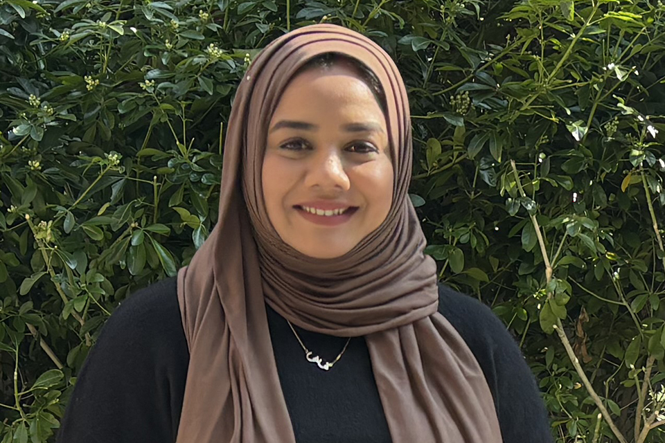 A woman wearing a brown hijab and a black top smiles while standing in front of green leafy bushes.