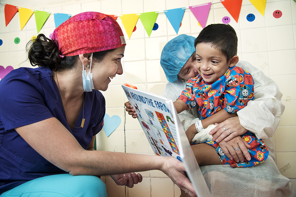 A nurse shows a colourful book to a smiling young boy in a hospital gown, who is sitting on an adult’s lap. The room is decorated with bright banners and paper hearts, creating a cheerful atmosphere.