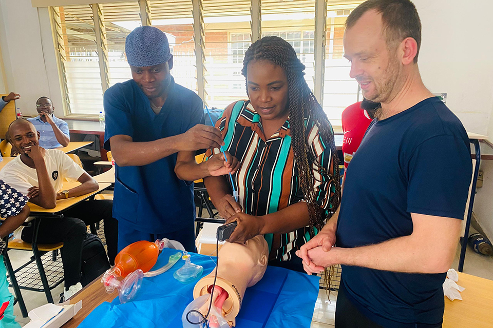 Three people practice a medical procedure on a mannequin arm in a classroom. One holds a syringe, another supports the arm, and the third observes. Medical equipment and students are visible in the background.
