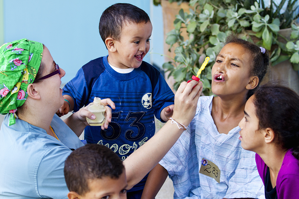 A woman blows bubbles with a group of smiling children outdoors; one girl is trying to catch a bubble while a young boy laughs excitedly. Lush green plants are in the background.